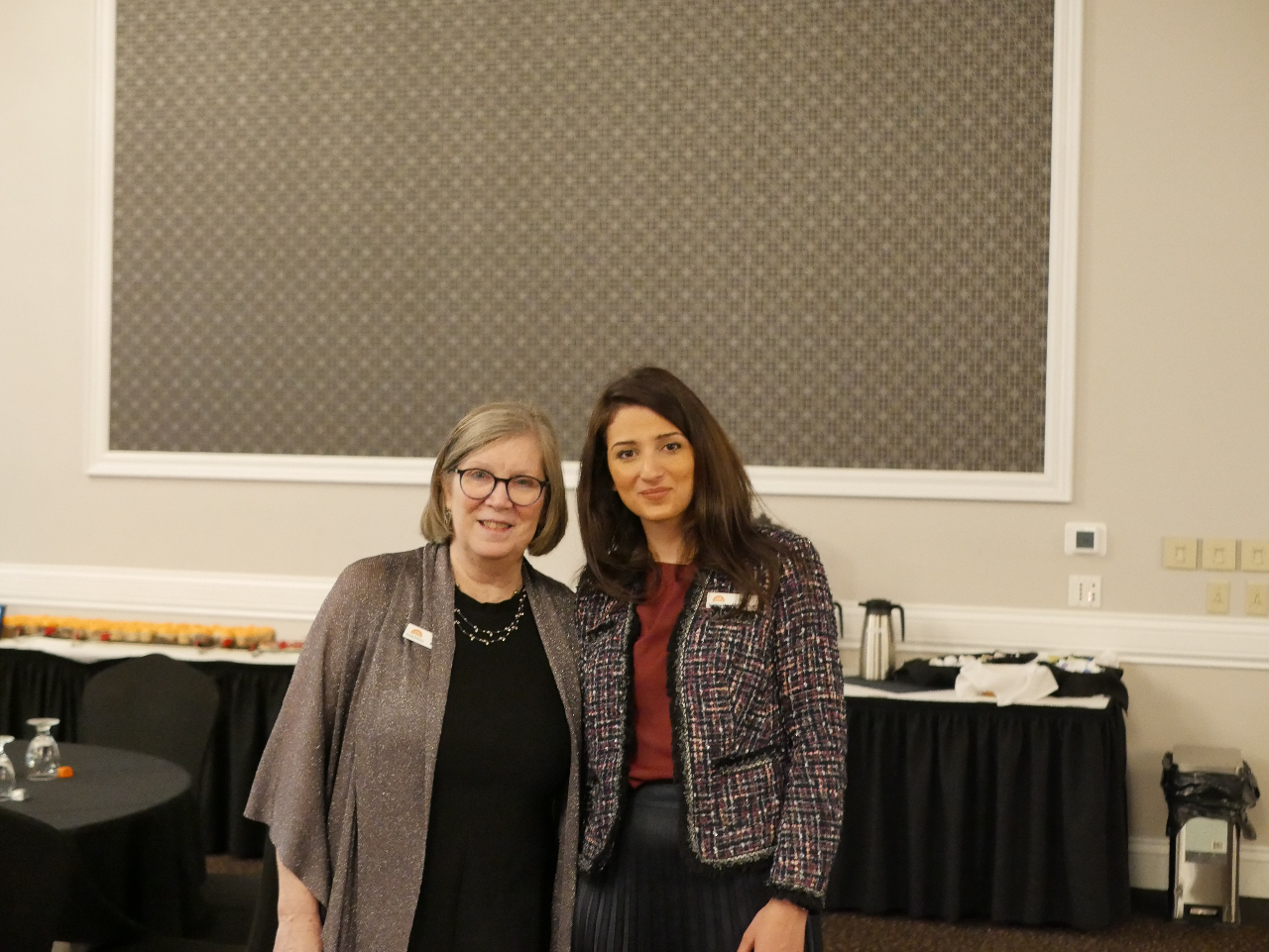 This Photo features WRFN Executive Director Sue Simpson and WRFN Fund Development Officer Oula Almadhoun, smiling side by side. Sue wears a grey shell with a black dress, and Oula wears a grey overcoat and has on a burgundy blouse.